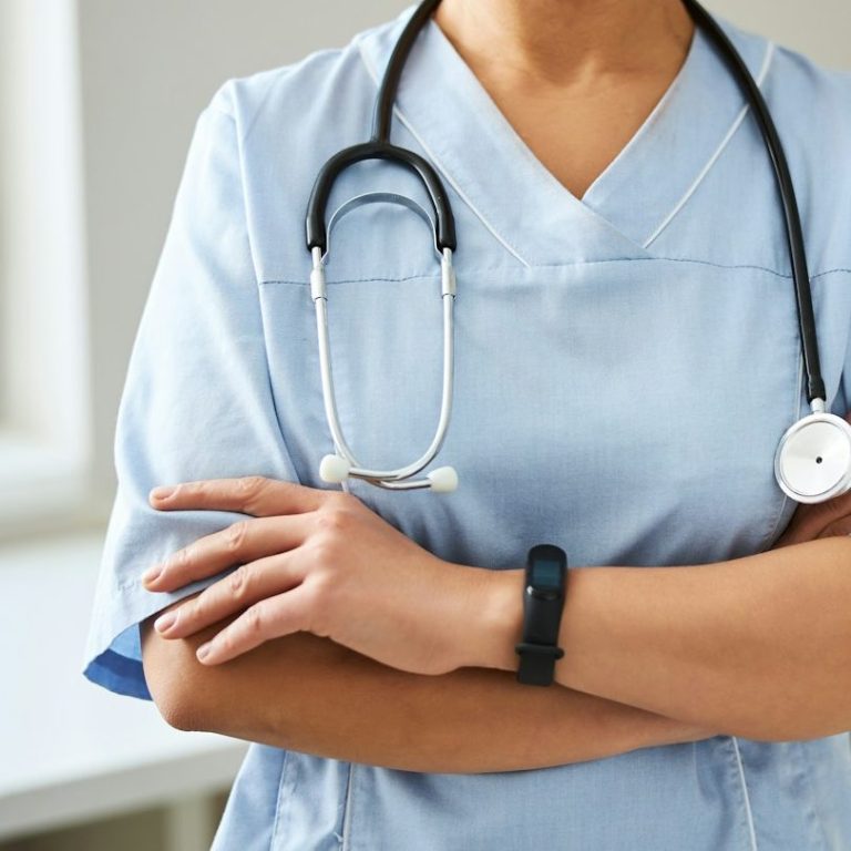 Eine Person in blauer Arztkleidung mit einem Stethoskop um den Hals und verschränkten Armen.