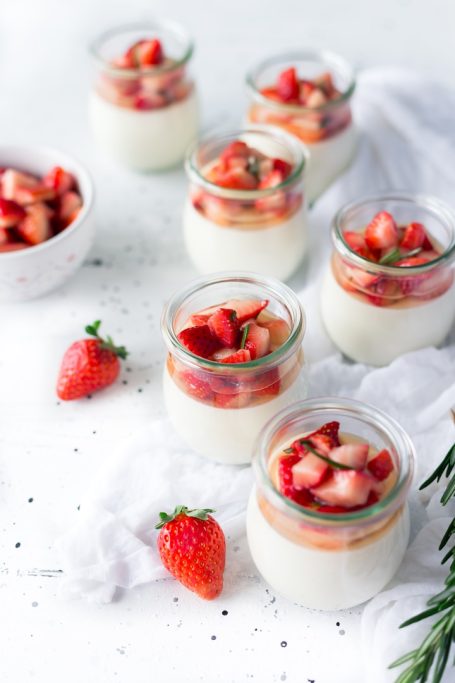 Sechs Gläser mit Pudding und frischen Erdbeeren, umgeben von Erdbeeren in einer Schale.